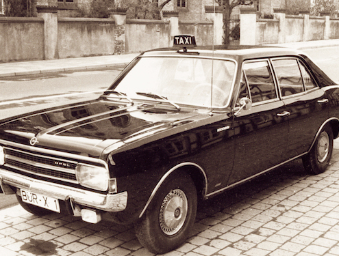 Ein altes Foto von einem Taxi. Das Auto ist ein schwarzer Opel und steht an der Strasse.